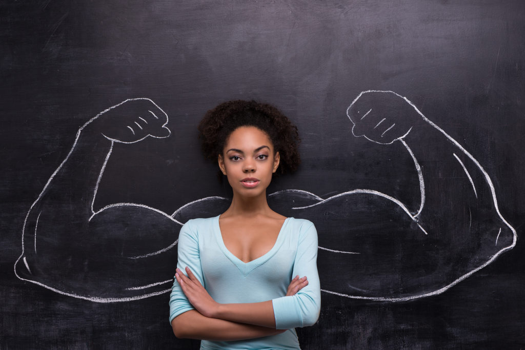 Funny picture of young afro-american woman on chalkboard background seriously looking at camera. Two strong muscular arms painted on chalkboard