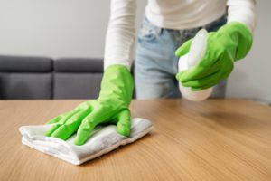 cropped shot of female hands in green rubber gloves hold detergent spray with rag and wipe wooden table from dust in living room , housework concept